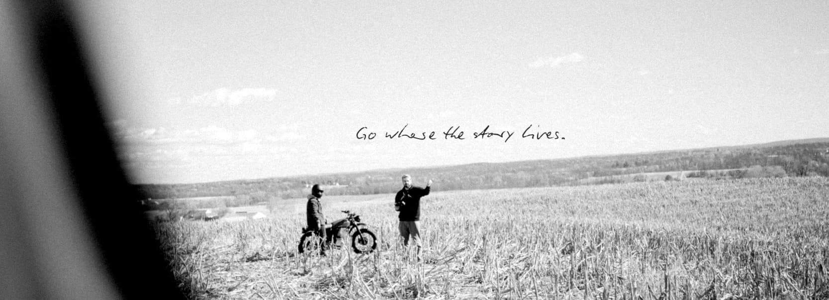 Two Aumen crew members standing with a dirt bike in an open field, captured through a car window. Hand-written across the sky: "Go where the story lives."