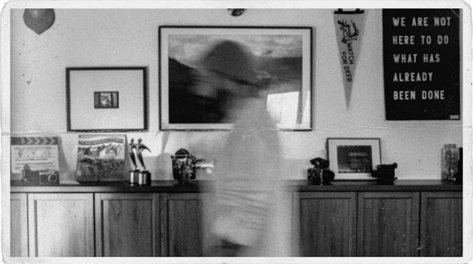 A vintage print with torn edges and a visible fold crease: a motion-blurred figure standing before a studio wall hung with framed artwork, a pennant reading "Watch for Deer," and a large text piece that reads: We are not here to do what has already been done.