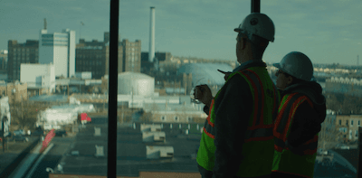 Two Kinsley Construction workers in hi-vis vests and hard hats look out a high window at the downtown skyline they're helping build.